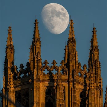 Photo of a nearly full moon and blue sky. The top of Yale University’s Harkness Tower in the foreground.